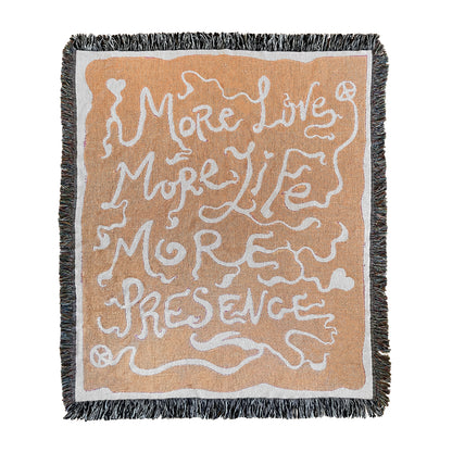 A rectangular woven blanket with a text pattern that reads 'MORE LOVE MORE LIFE MORE PRESENCE' on an orange background, with a grey fringe border.