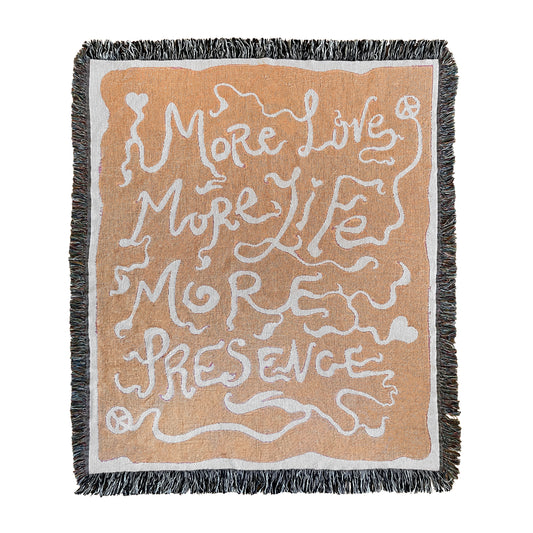 A rectangular woven blanket with a text pattern that reads 'MORE LOVE MORE LIFE MORE PRESENCE' on an orange background, with a grey fringe border.